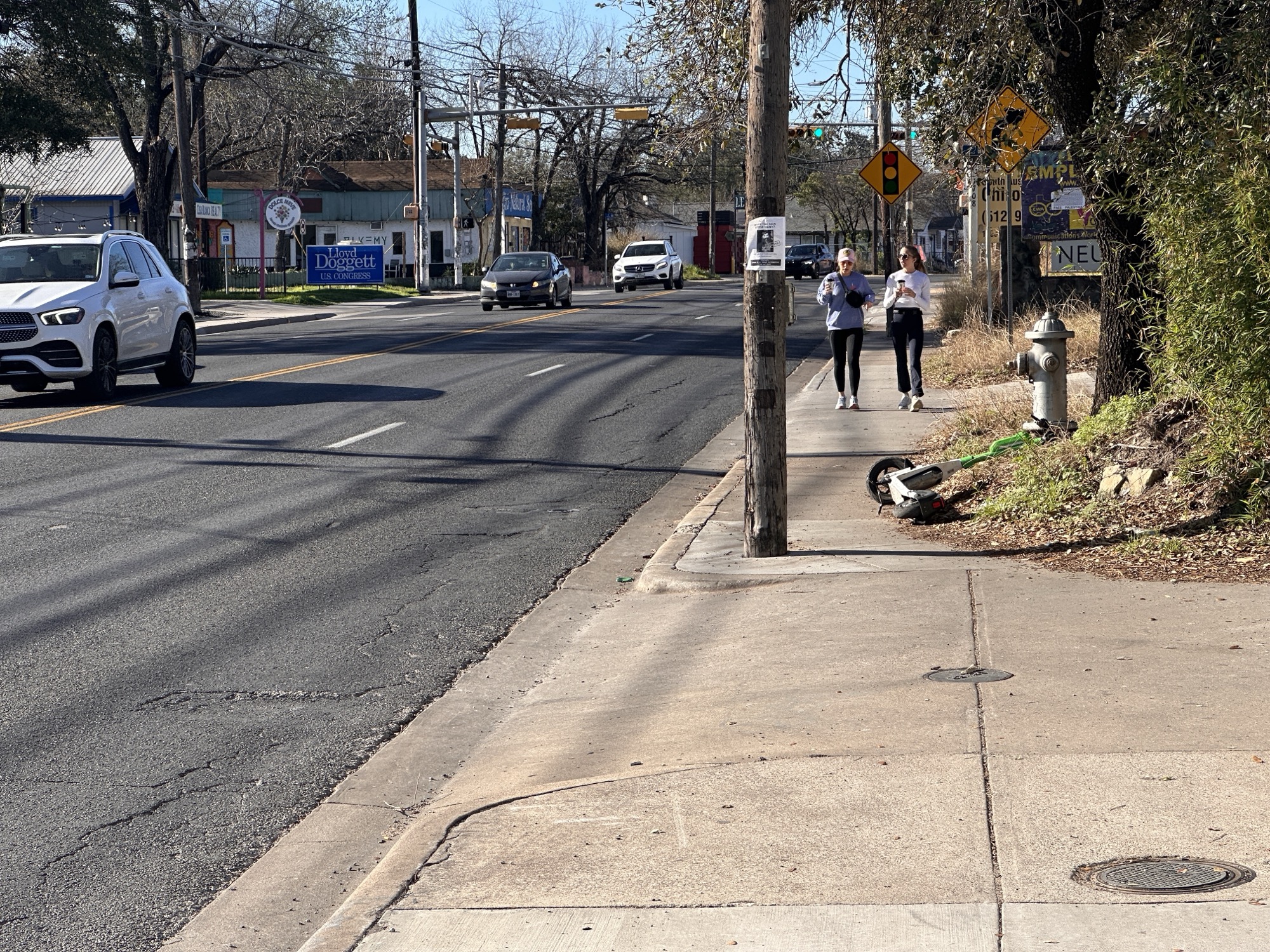 Two people walking on a narrow South First sidewalk beside four-lane traffic, utility poles, and roadside signs